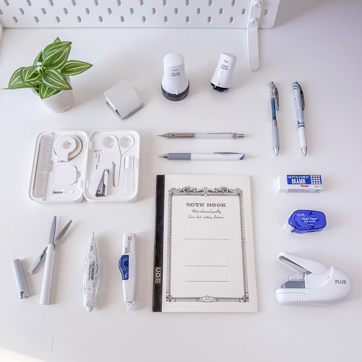 Flat lay of a decorative notebook surrounded by pens pencils erasers correction tape scissors a stapler and other stationery on a white desk with a small plant