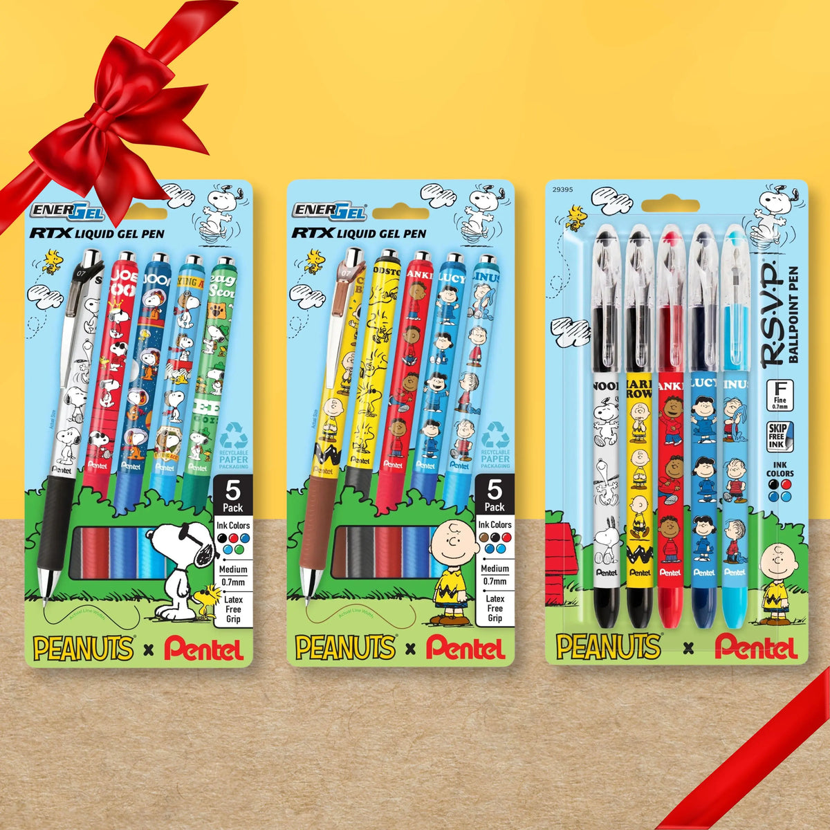 Three Peanuts and Pentel packages of colorful gel pens and ballpoint pens arranged on a yellow and wood background with a red ribbon bow and gift wrap ribbon
