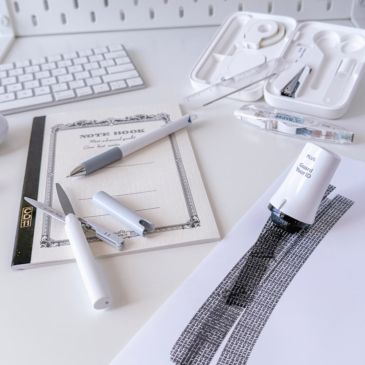 White desk with a notebook, pens, pencil, ruler, eraser, correction tape, keyboard, and printed paper arranged for writing and drawing.