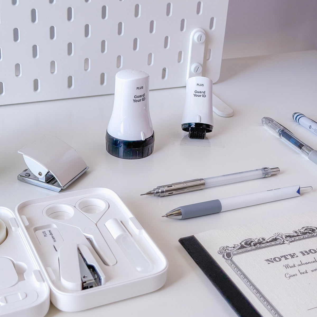 White desk arrangement with Guard Your ID stamp tools pens a stapler pencil case and a decorative notebook on a clean workspace