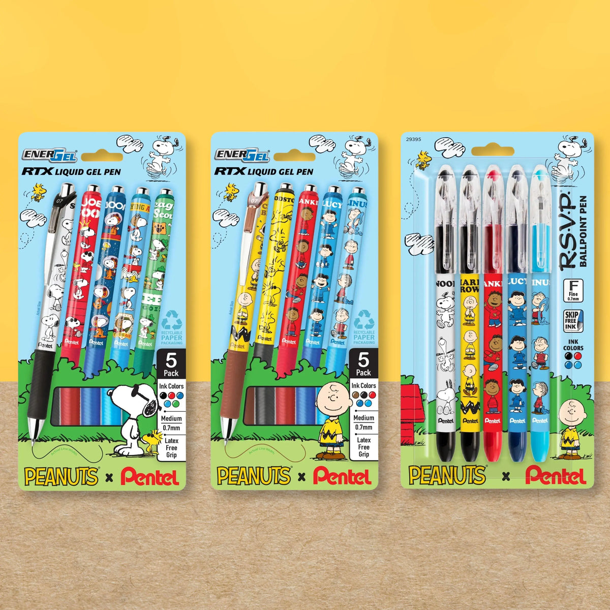 Three colorful Peanuts themed Pentel pen packages displayed on a yellow and wood grain background, including liquid gel and ballpoint pens in assorted colors