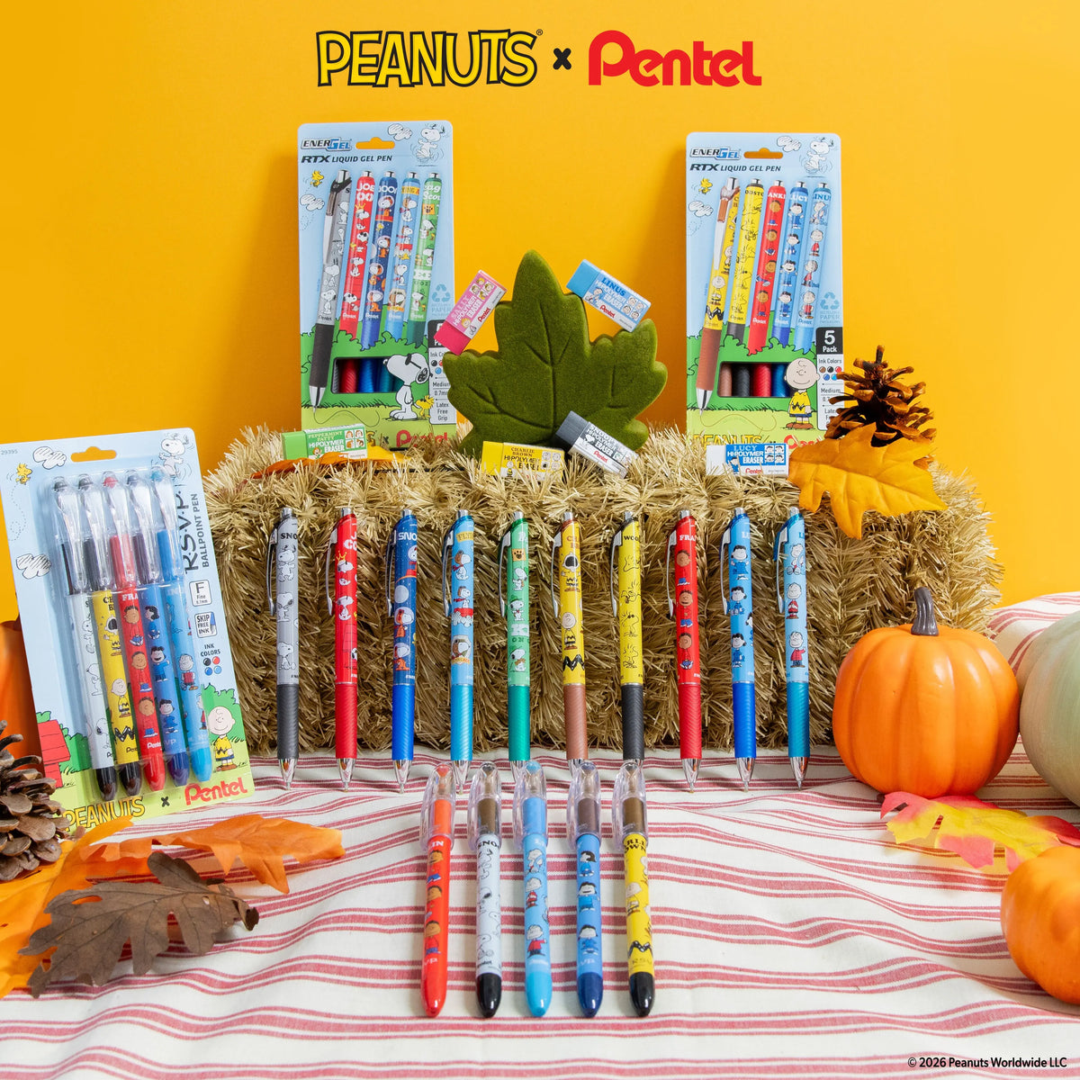 Peanuts and Pentel themed display of colorful gel pens arranged on a striped tablecloth with autumn leaves, pumpkins, pinecones, and packaged pens in the background