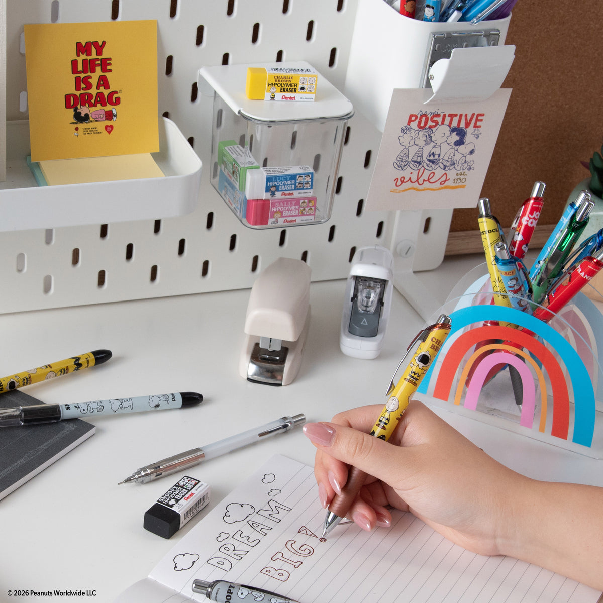 Hand using a yellow pencil to draw on lined paper beside pens mechanical pencils an eraser and colorful desk organizers.