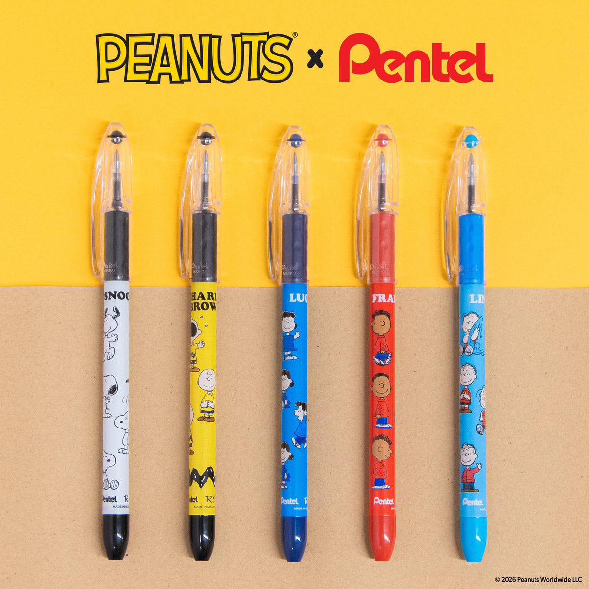 Five colorful Peanuts and Pentel pens with character designs, clear caps, and black grips arranged on a yellow and kraft paper background