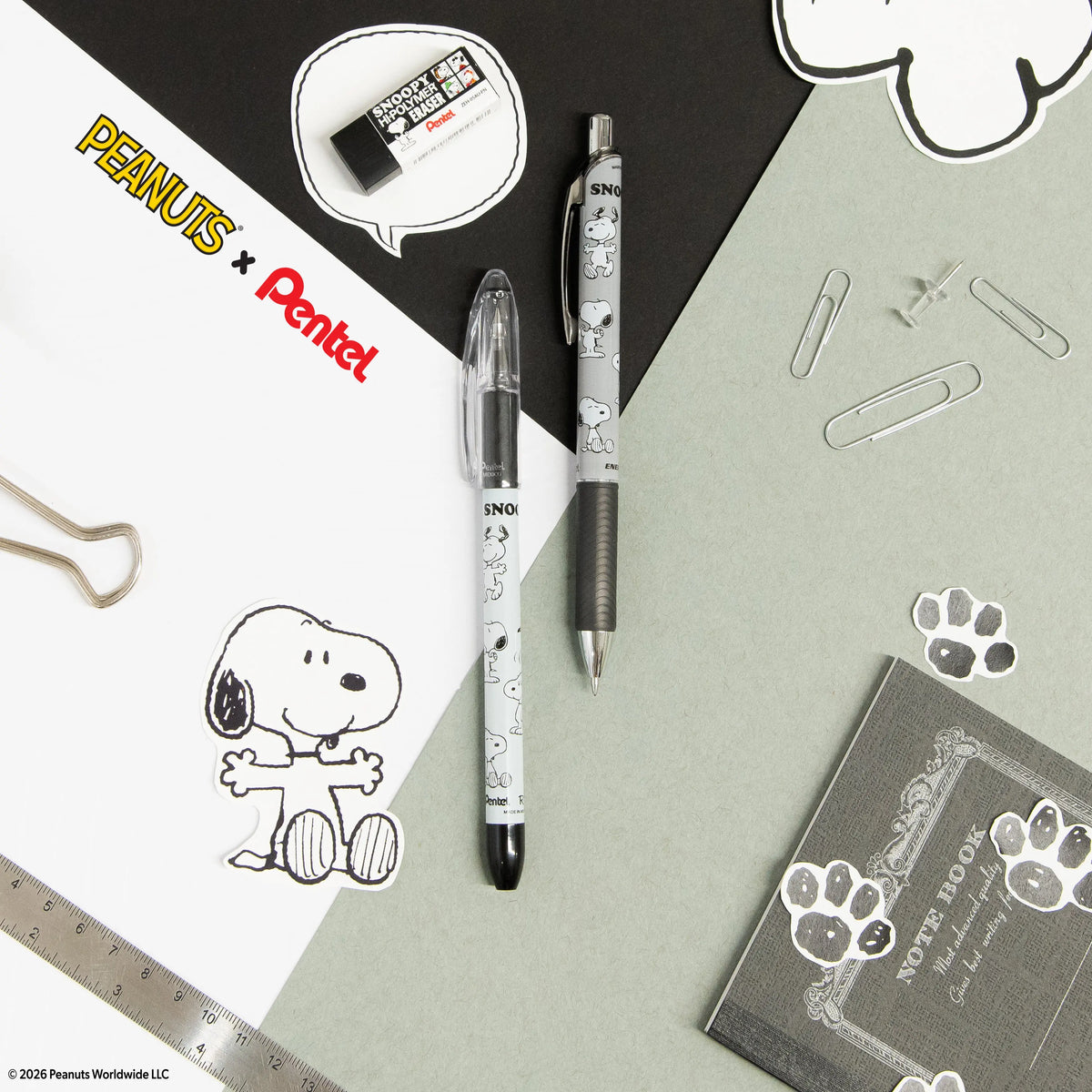 Snoopy themed Pentel pens arranged on paper with a ruler, paper clips, eraser, notebook, and Peanuts artwork visible around them