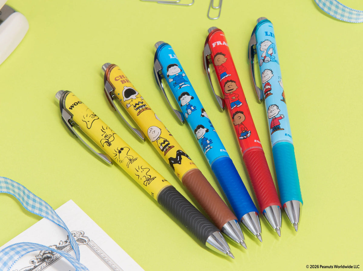Five Peanuts themed Pentel EnerGel gel pens in yellow blue red and turquoise with character decorated barrels arranged on a lime green surface