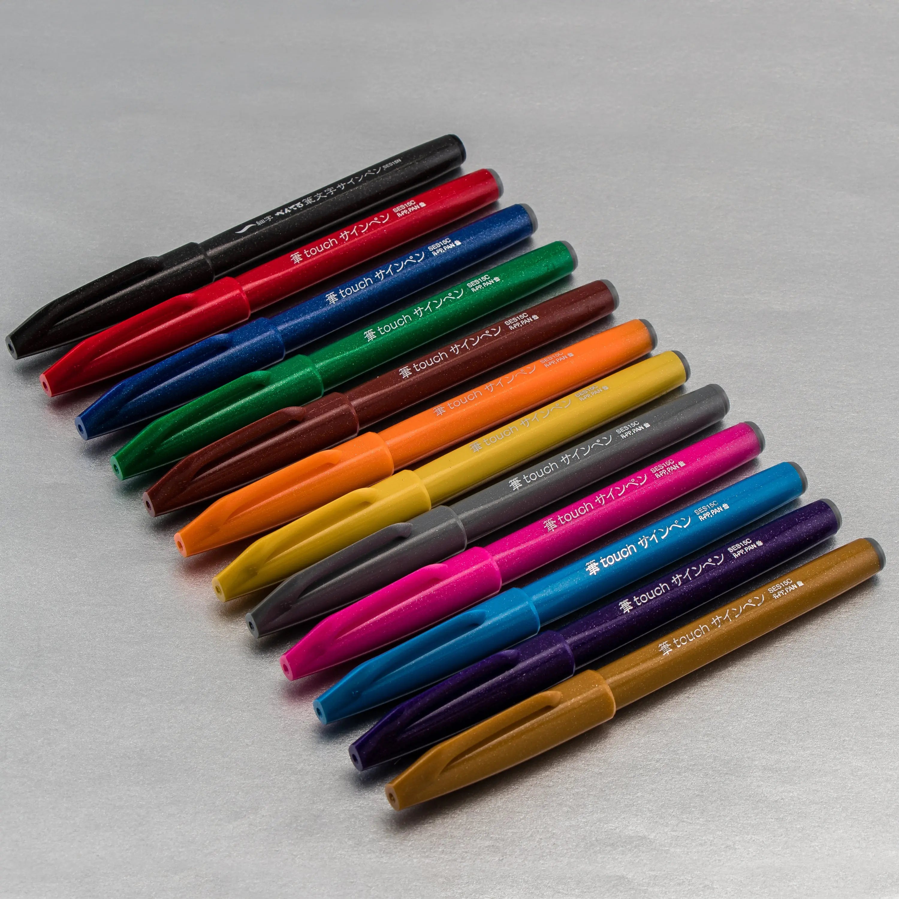 Pentel Arts brush tip pens in assorted vibrant colors arranged on a gray surface for expressive lettering calligraphy illustration and creative art projects