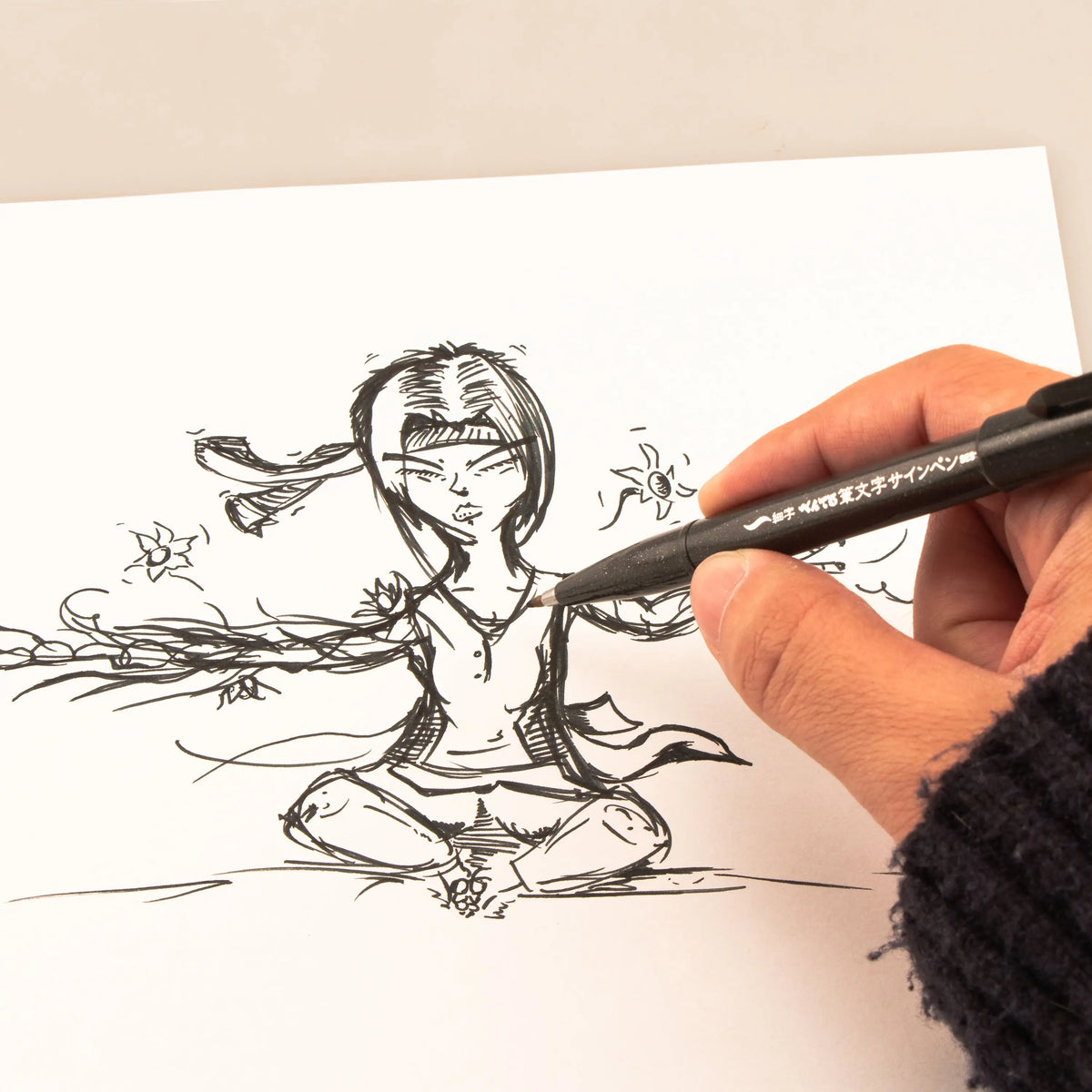 Hand using a black brush tip pen to sketch a detailed seated figure with flowing lines on white paper for expressive artwork and calligraphy practice