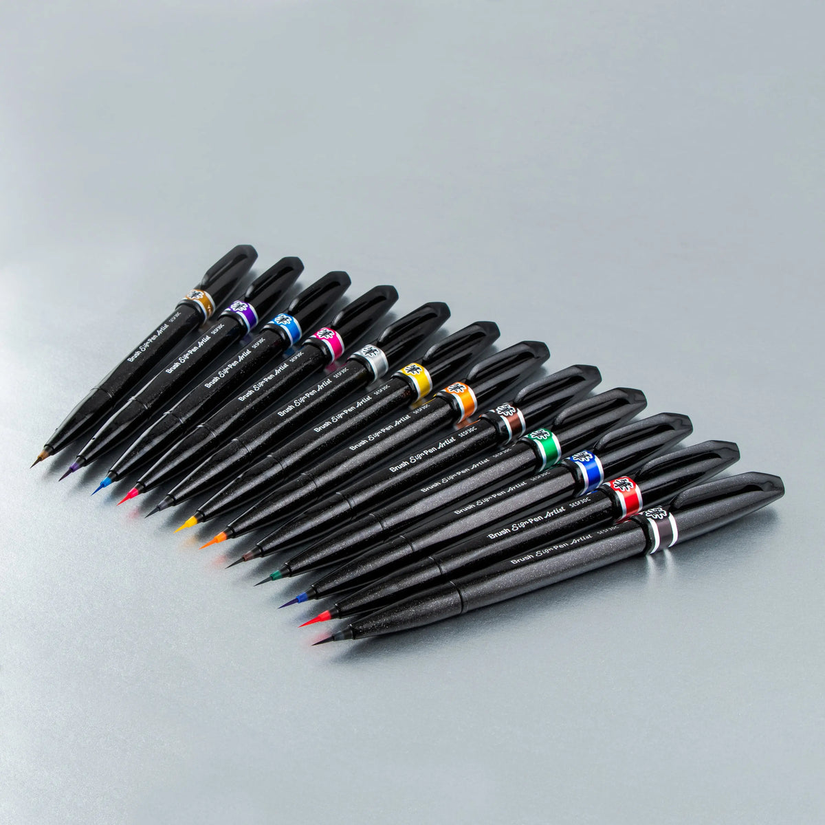 Pentel Arts micro brush pens with black barrels and assorted ink colors arranged in a fan on a light surface for detailed drawing and lettering
