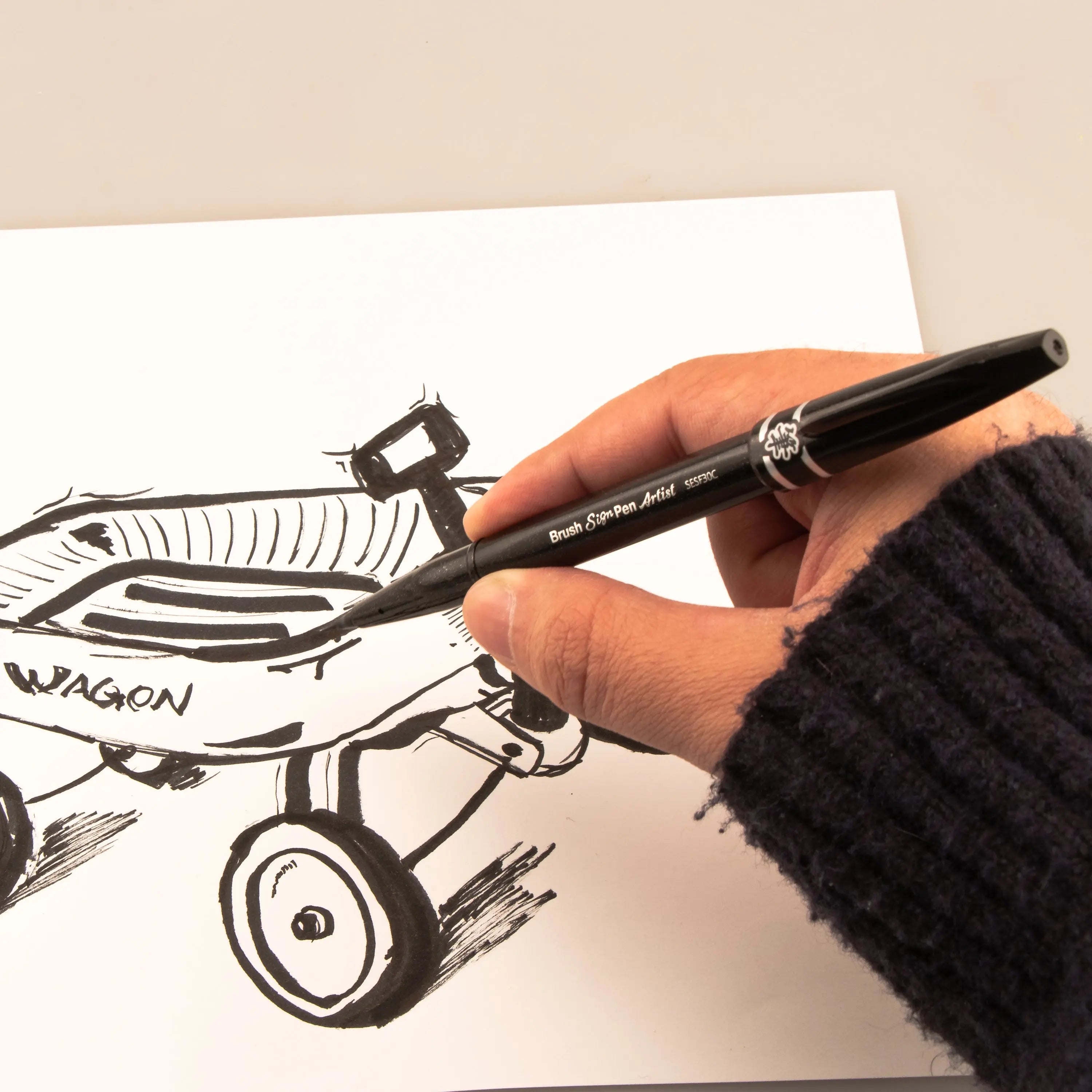 Hand using a black micro brush pen to add bold ink lines and fine details to a whimsical vehicle sketch on white paper