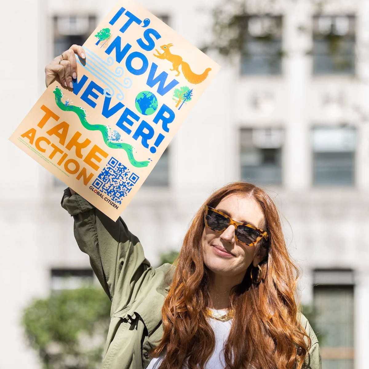 Woman wearing sunglasses holds an illustrated sign reading ITS NOW OR NEVER TAKE ACTION with a QR code outdoors near a building.