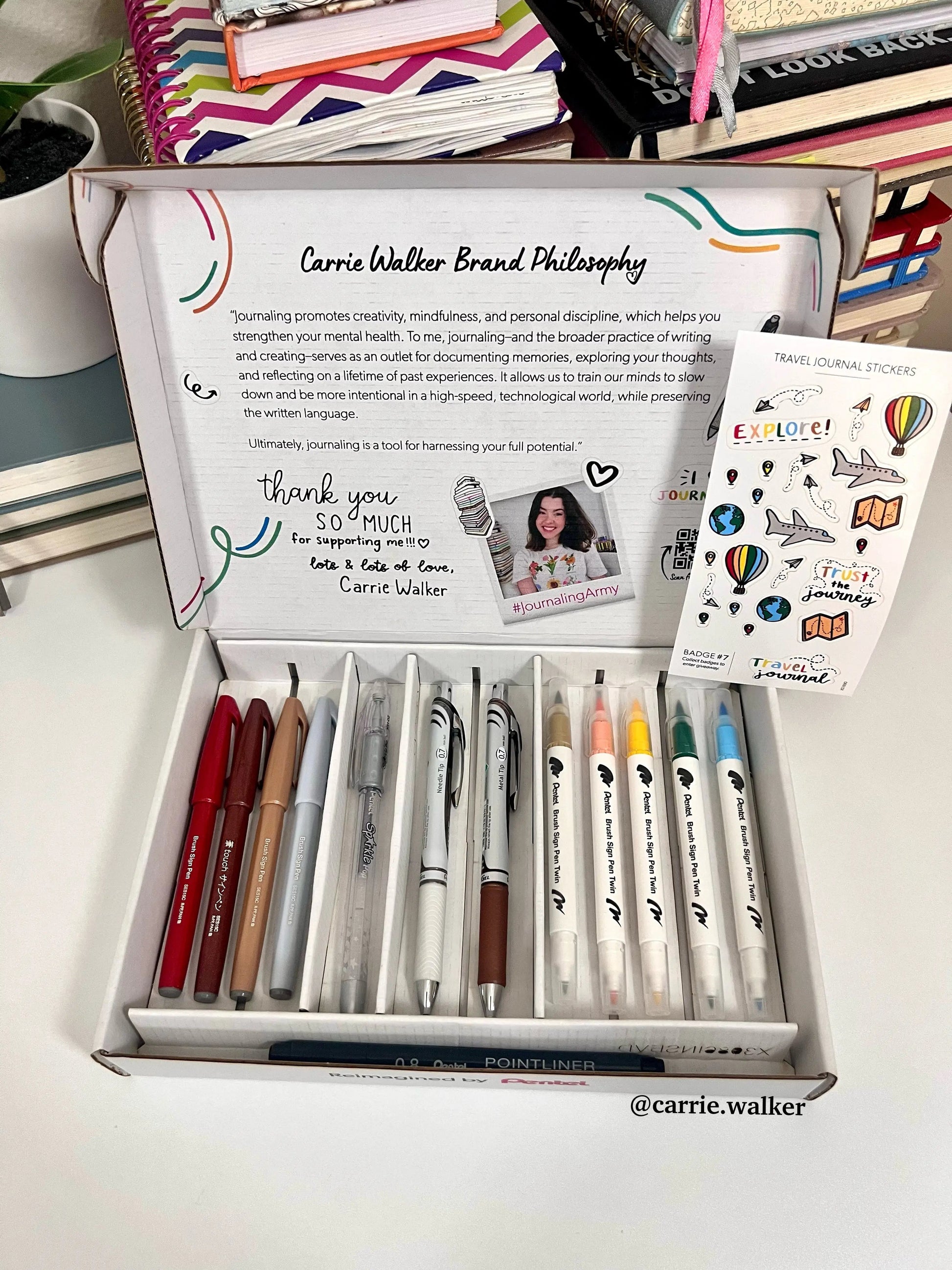 Open box of colorful gel pens, markers, and mechanical pencils arranged neatly with a set of travel-themed journaling stickers, promoting mindful creativity and premium writing tools.