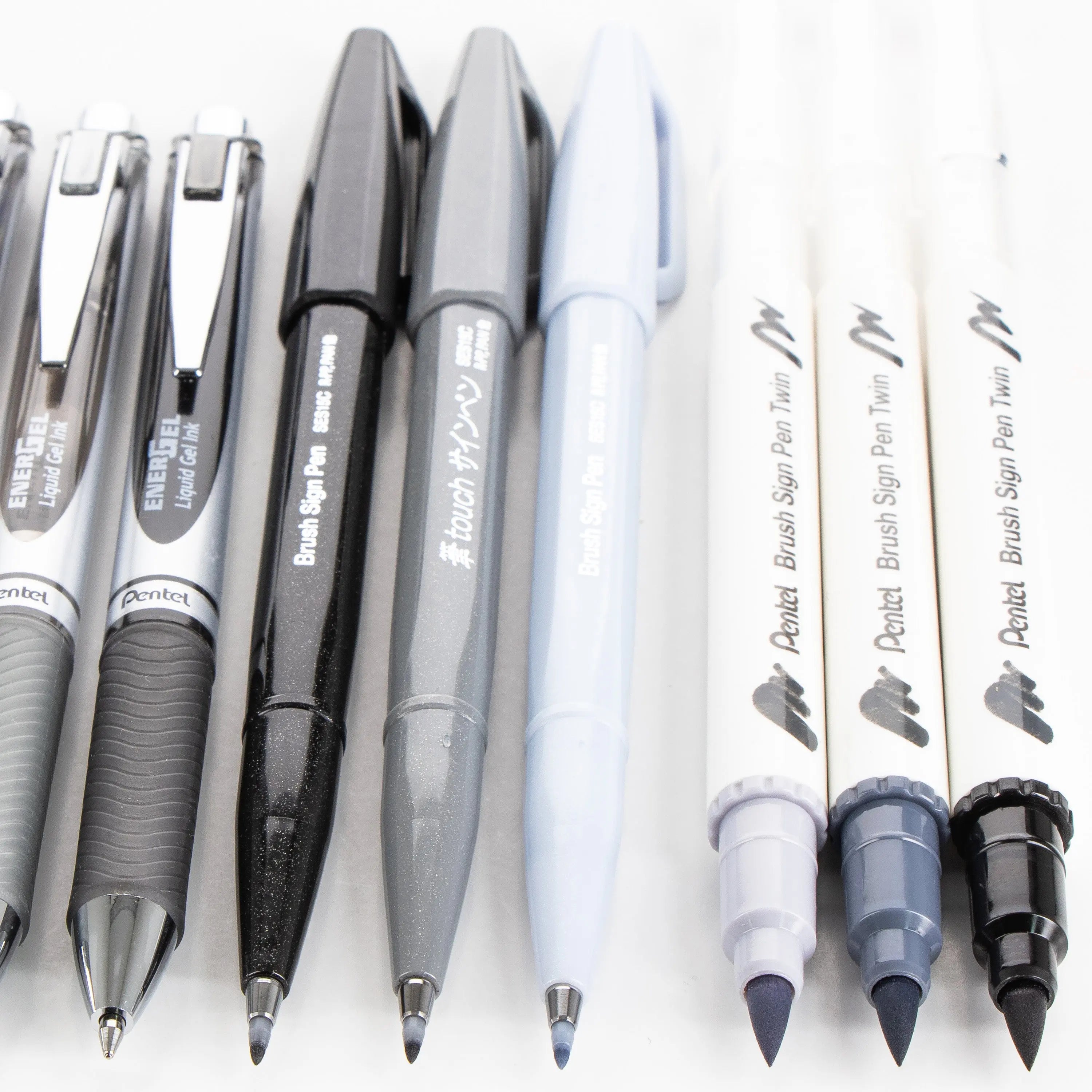 Close-up of a variety of high-quality gel pens and brush pens arranged in a row with black, grey, light blue, and white barrels, showcasing smooth ink tips and ergonomic grips for premium writing and creative expression.